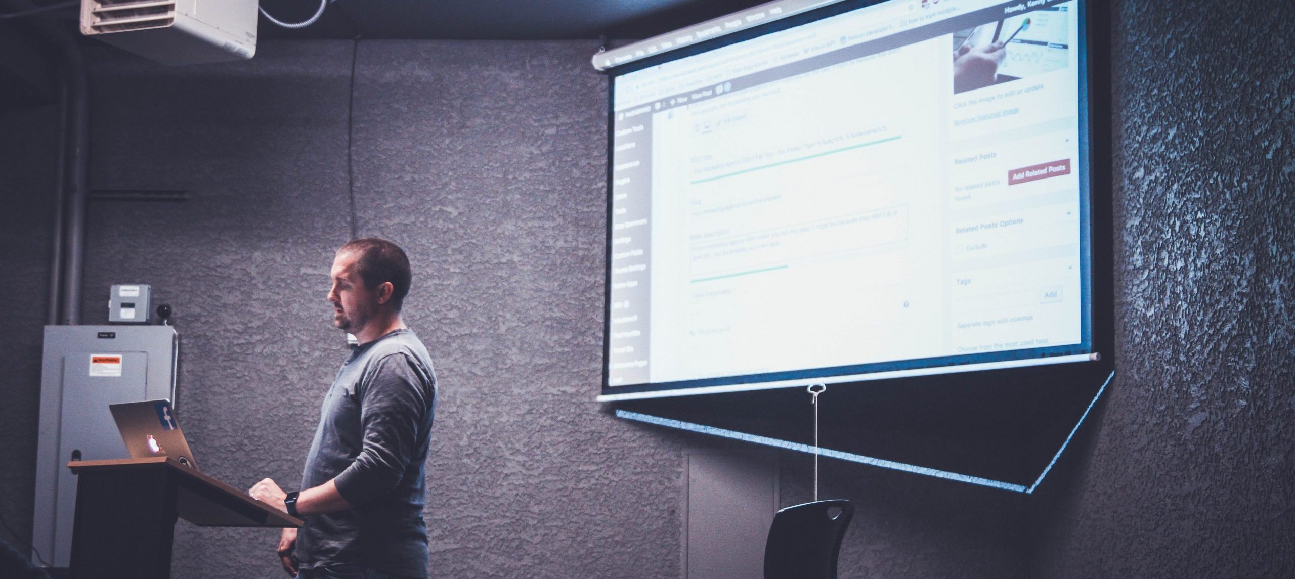 Personne se tenant devant un ordinateur portable sur un pupitre et présentant un cours PowerPoint sur un écran de projection.