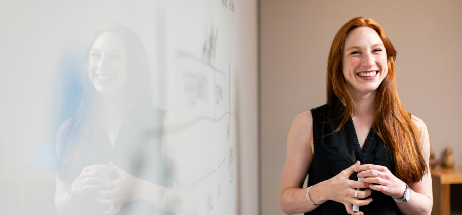 A person with long red hair smiles, standing beside a reflective surface in a room. They're wearing a sleeveless black top, pondering ways to make online courses more interactive.