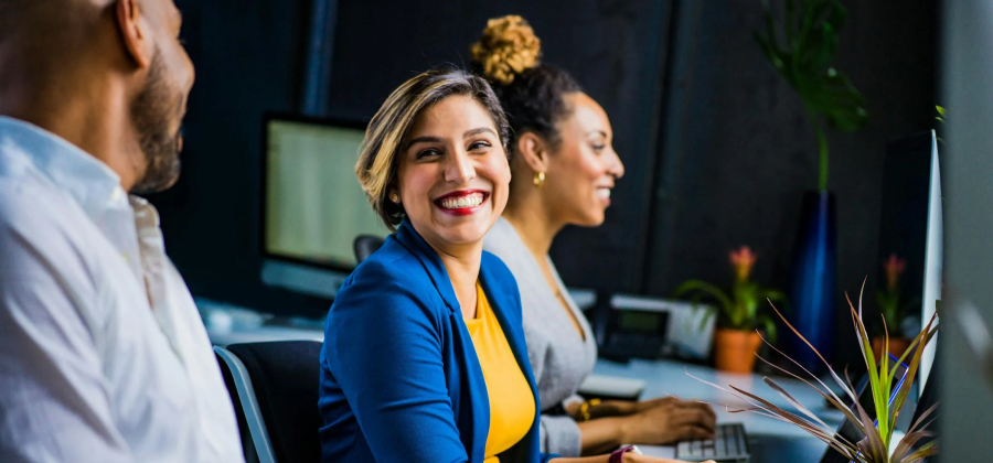 Three people sit at desks in an office setting, engaging with a corporate training platform. Two women smile warmly, while a man turns to the side in concentration. A lush plant graces the foreground, adding a touch of nature to the professional environment.